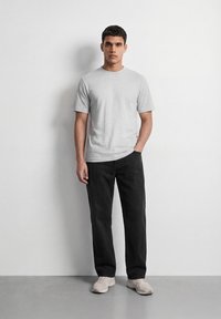 Νεαρός άνδρας στέκεται μπροστά σε λευκό τοίχο φορώντας γκρι κοντομάνικο μπλουζάκι, μαύρο παντελόνι και ανοιχτόχρωμα αθλητικά παπούτσια.