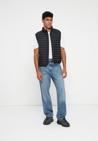 Sort polstret vest med høj krave, båret over en hvid t-shirt, parret med lyseblå jeans og sorte sko.