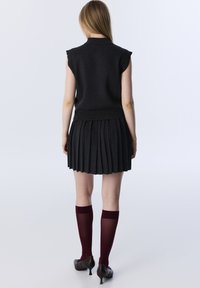 Dunkelgraues ärmelloses Oberteil kombiniert mit einem plissierten Rock, burgunderroten Kniestrümpfen und schwarzen hohen Schuhen. Glatte Textur mit minimalistischem Design.