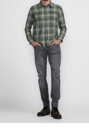 Homme portant une chemise à carreaux verte à boutons, un jean gris, des bottes marron à lacets, debout les mains dans les poches devant un fond uni.