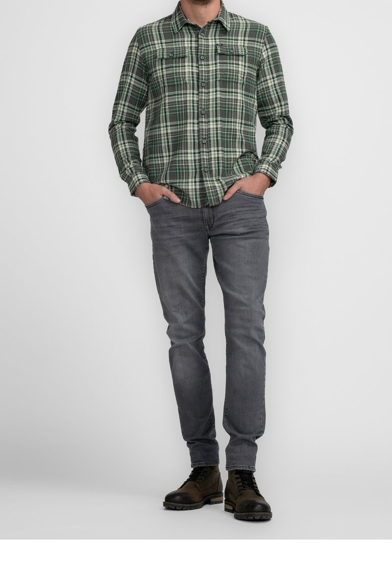 Homme portant une chemise à carreaux verte à boutons, un jean gris, des bottes marron à lacets, debout les mains dans les poches devant un fond uni.