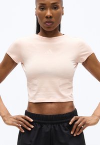 Αθλητική γυναίκα που στέκεται με τα χέρια στους γοφούς, φορώντας ανοιχτό ροζ κοντομάνικη μπλούζα και μαύρα σορτς με ελαστική μέση, πάνω σε λευκό φόντο.