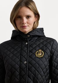 Veste matelassée noire avec capuche, boutons-pression argentés et logo brodé doré sur la poitrine gauche. Texture lisse avec un motif en losanges.