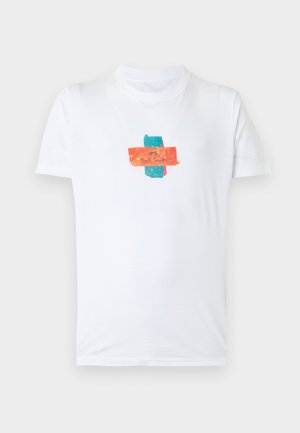 Jeune homme aux cheveux bouclés portant un t-shirt graphique blanc, un pantalon noir et des baskets blanches, debout les mains dans les poches sur un fond uni.