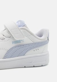 Baskets blanches avec des accents argentés, semelle en caoutchouc texturée, tige en cuir lisse et bande velcro. Présente une rayure bleu clair et un logo.