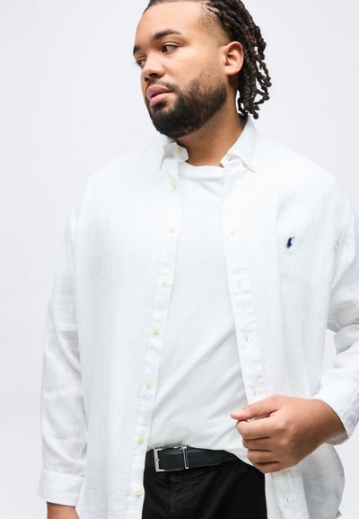 Άνδρας με πλεξούδες στα μαλλιά και γένια, φορώντας λευκό πουκάμισο με κουμπιά πάνω από λευκό T-shirt, που ρυθμίζει το πουκάμισο με το δεξί χέρι, μπροστά σε απλό φόντο.