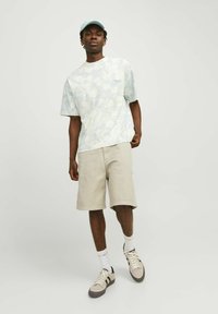 T-shirt corto a maniche corte blu chiaro marmorizzato, pantaloni beige, calze bianche e sneakers beige/grigie indossati da un modello maschile in posizione eretta.