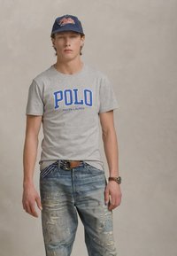 Tricou gri din bumbac cu logo-ul "Polo" albastru, combinat cu blugi albaștri distruși, care prezintă uzură vizibilă și o curea din piele maro. Include o șapcă închisă la culoare.