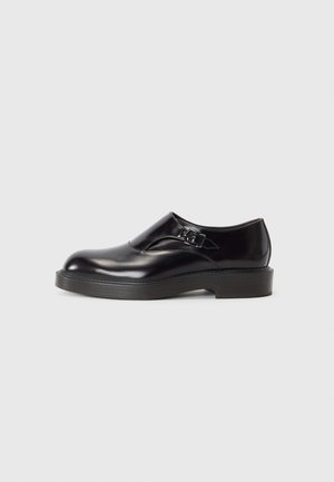 Black leather shoe with a rounded toe, low black heel, and a decorative strap. Smooth texture with a shiny finish.