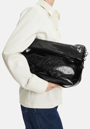 Person wearing cream shirt and blue jeans holding a large black shiny leather handbag under their arm.
