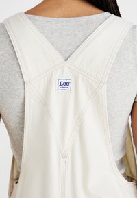 Κρέμα βαμβακερή φόρμα με ροζ ραφές, με σχέδιο X-back και μπλε ετικέτα Lee. Γκρι ριχτό πουκάμισο από κάτω.