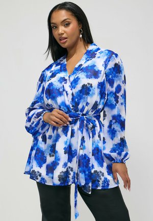 Mujer con blusa cruzada de manga larga azul y negra con estampado floral y cinturón en la cintura, combinada con pantalones negros, posando contra un fondo liso.