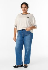 Top de punto oversize color beige claro con detalles de rayas negras, combinado con vaqueros azules de tiro alto y pierna recta, y zapatos planos negros.