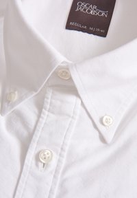 Camicia bianca in cotone con colletto button-down, quattro bottoni di chiusura e un'etichetta in tessuto contrassegnata "OSCAR JACOBSON." Texture morbida.