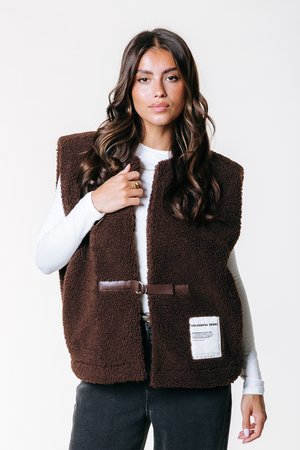 Gilet in fleece marrone con colletto alto, cintura simile alla pelle e tasca applicata con testo bianco. Indossato sopra una camicia bianca a maniche lunghe.