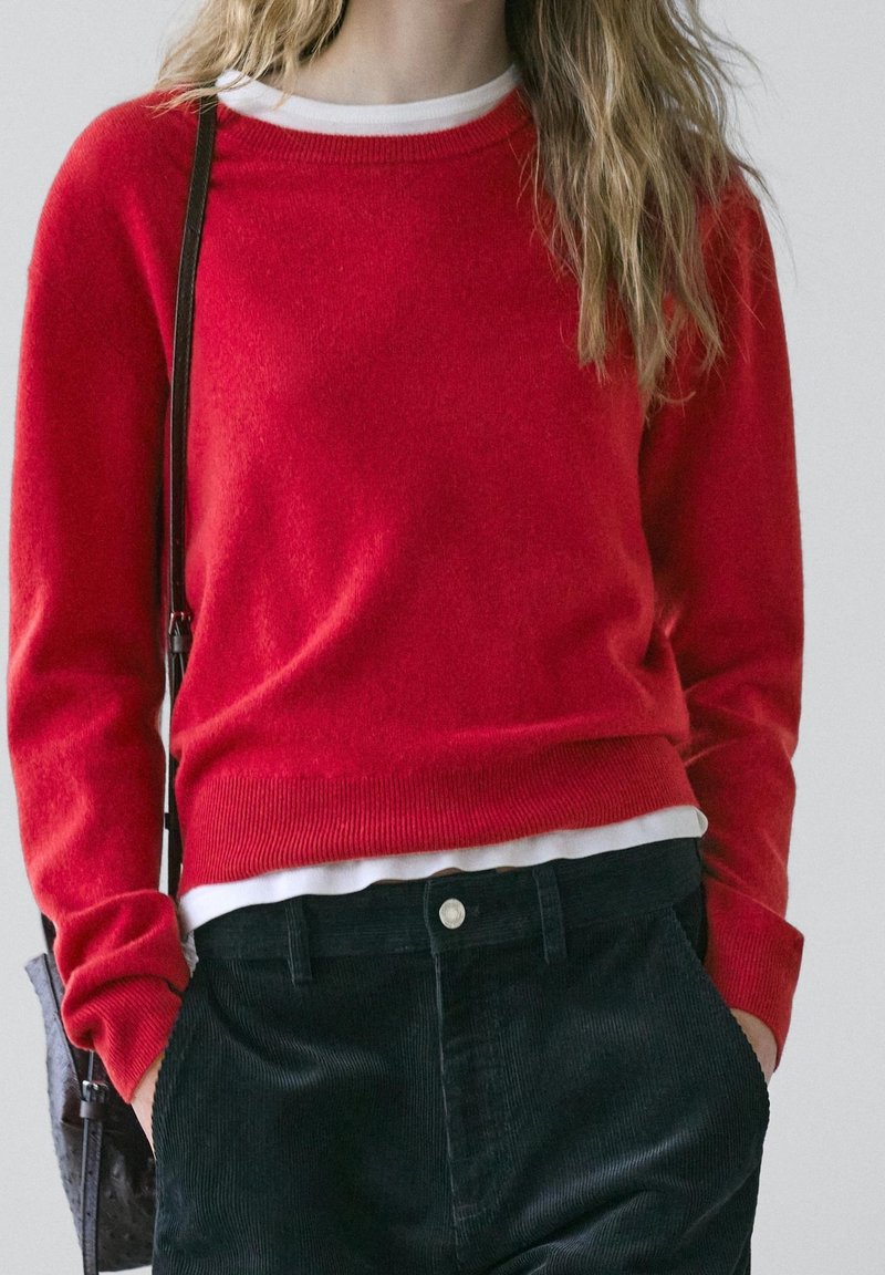 Femme portant un pull rouge sur une chemise blanche, un pantalon en velours côtelé noir, les mains dans les poches et une bandoulière de sac à main noire visible.