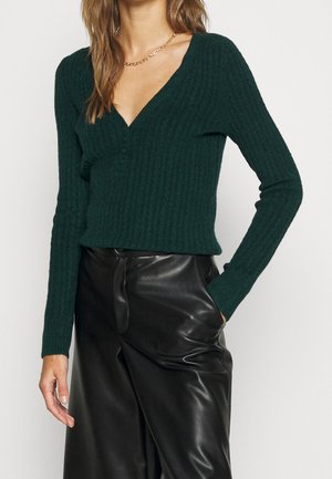 Femme portant un pull en maille côtelée vert foncé avec des boutons et un pantalon en cuir noir, main dans la poche, sur un fond uni.