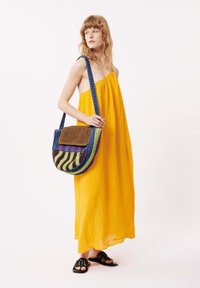 Gelbes ärmelloses Maxikleid aus strukturiertem Stoff; mehrfarbige, gewebte Schultertasche mit horizontalen Streifen und einem braunen Deckel; schwarze Sandalen.