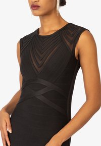 Schwarzes Kleid mit einer figurbetonten Silhouette, transparenten Einsätzen und strukturierten vertikalen Streifen. Das Design umfasst geschichtete geometrische Akzente.