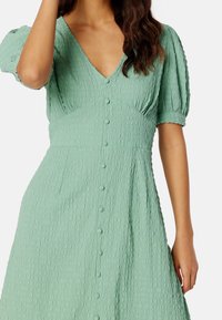 Ljust grönt puffärmad klänning med V-ringning, texturerad tyg och knapprad fram. A-linjeform med två sidofickor.