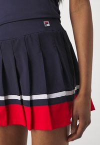 Mörkblå tennisplisserad kjol med röda och vita ränder vid fållen. Har en liten märkt etikett på midjebandet. Slätt, lätt tyg.