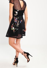 Svart klänning med korta meshärmar och blommig broderi i olika färger. Kombinerad med svarta klackar med remmar och en nyckelhålsöppning bak.