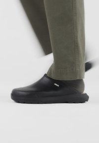 Chaussures noires à enfiler avec une tige texturée et une semelle robuste, associées à un pantalon vert olive, mettant en valeur un design simple et fonctionnel.