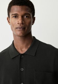 Camiseta polo negra con cuello corto, tres botones negros y un bolsillo en el pecho. La tela parece suave y de punto, con una textura uniforme.