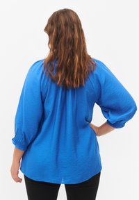 Blusa ligera azul con un acabado texturizado, mangas abullonadas de tres cuartos y un dobladillo ligeramente curvado. Llevada sobre pantalones negros.