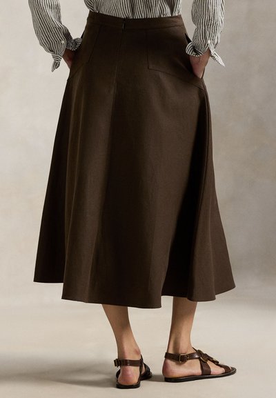 Καφέ, μακριά φούστα σε ροή με εφαρμοστό ζωνάκι, πλαϊνές τσέπες και μαλακή υφή, συνδυασμένη με καφέ σανδάλια με λουράκια.