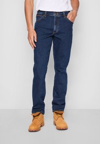 Homme portant un jean bleu foncé, des bottes de travail marron clair et une chemise blanche, debout avec une main dans la poche devant un fond uni.