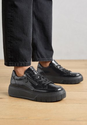 Personne portant des baskets en cuir verni noir avec des semelles épaisses et des fermetures éclair latérales, debout sur un sol en bois avec un pantalon noir.