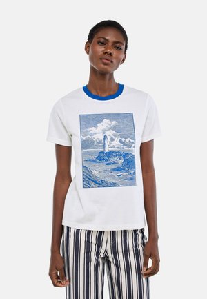 Femme portant un T-shirt blanc avec un col bleu et un motif de phare bleu, associé à un pantalon rayé vertical noir et blanc.