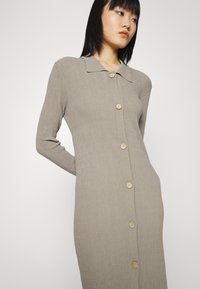 Vestido de manga longa com nervuras, de botão, na cor taupe claro, com colarinho; apresenta botões de madeira na frente e uma silhueta ajustada.