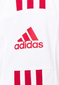 Vit sporttröja med röd Adidas-logga och tre vertikala röda ränder på vänster sida. Tillverkad av ett slätt, lätt material.