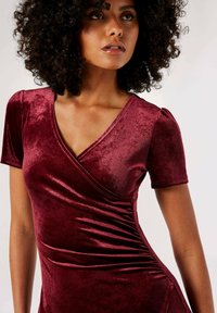 Burgunderfarbenes Samtkleid mit kurzen Ärmeln, V-Ausschnitt und gerafften Seitendetails. Weiche Textur und taillierte Silhouette.