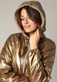 Veste rembourrée en or métallique avec une capuche doublée de fausse fourrure, fermeture éclair à l'avant et poignets élastiques. Texture lisse, design volumineux.