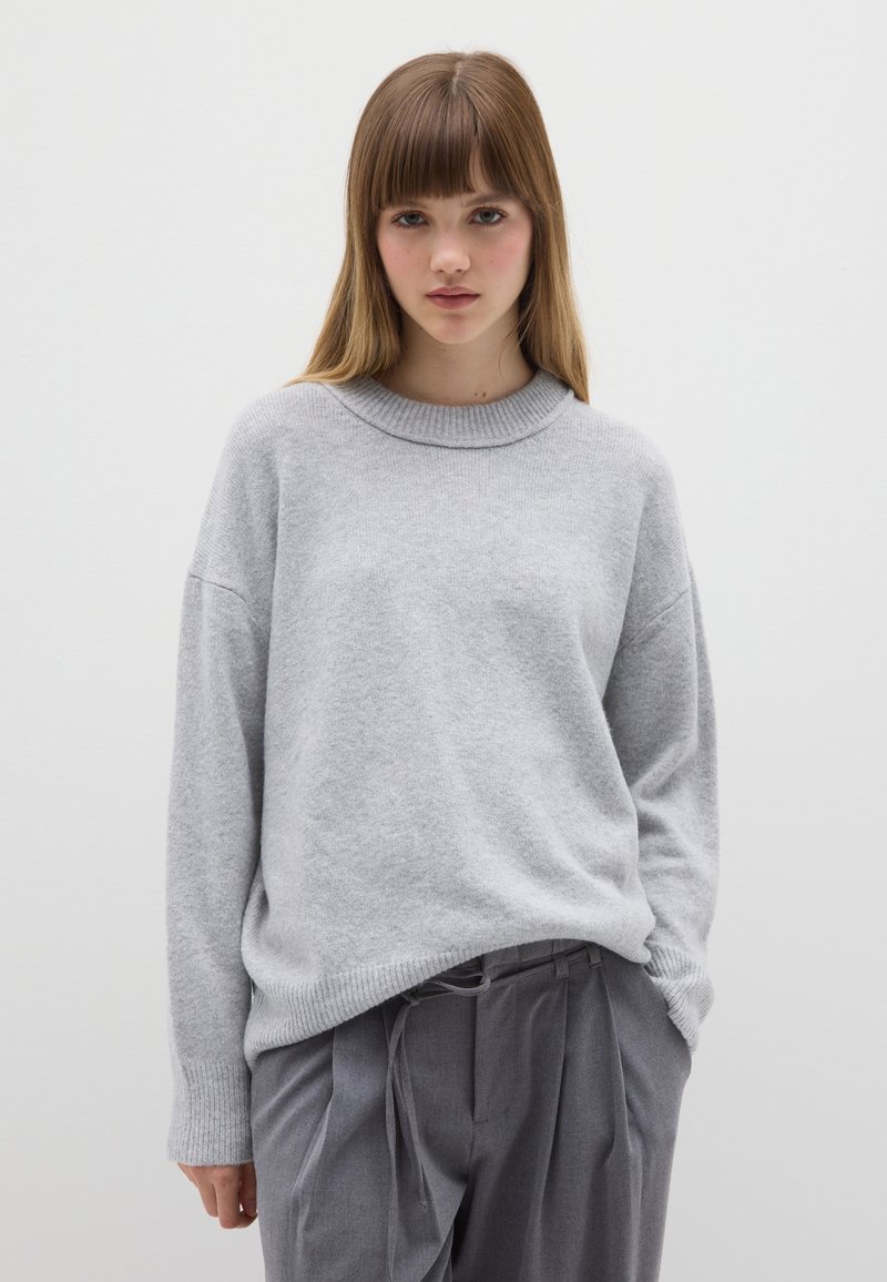 Hellgrauer, übergroßer Pullover mit geripptem Rundhalsausschnitt und langen Ärmeln. Weiche Textur, minimalistisches Design, kombiniert mit grauer Hose.