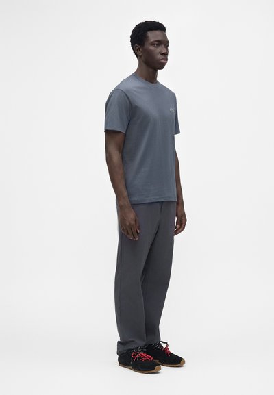 Homme debout portant un t-shirt gris, un pantalon gris foncé et des baskets noires avec des lacets rouges sur un fond blanc uni.