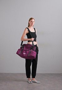 Lilla duffelveske med to svarte stropper og en toppklaff. Glatt materiale med en subtil glans. Robust design, romslig og funksjonell.