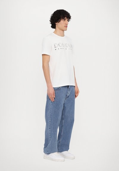T-shirt en coton blanc avec "DONDU" imprimé en argent, associé à un jean baggy bleu clair et des baskets blanches. Le mannequin se tient de côté.