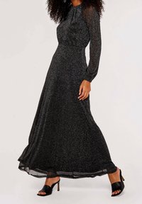 Musta, lattiaan asti ulottuva mekko, jossa pitkät läpikuultavat hihat, kimaltava pinta ja kerätty pääntie, yhdistettynä avoimilla varpailla oleviin mustiin korkokenkiin.