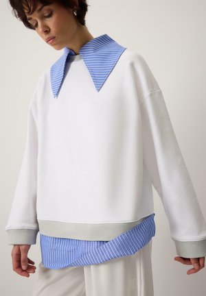 Donna che indossa un maglione bianco oversize con polsini grigi e un grande colletto e orlo a righe blu, sopra pantaloni bianchi.
