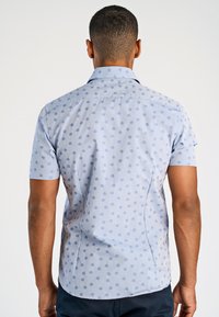 Camicia a maniche corte, di colore azzurro con un motivo di pois blu scuro. Presenta un colletto tradizionale e un orlo arcuato.