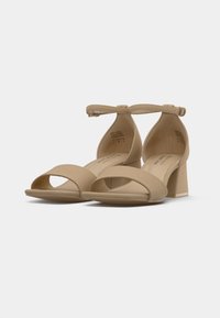 Sandales à talons beiges avec une bride à la cheville, bout ouvert et design avant arrondi. Elles sont dotées d'un matériau texturé et doux ainsi que d'un talon bloc solide.