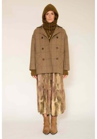 Manteau croisé à carreaux bruns avec de grandes poches, superposé sur un pull en maille brun et une longue jupe tie-dye. Bottes de cowboy brunes.