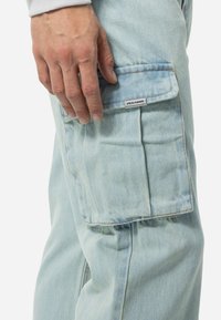 Lys blå denim cargo bukser med en sidelomme, kontraststikning og et mærkat på lommen. Nærbillede af en hånd, der hviler på bukserne.