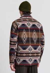 Fleece-Jacke mit mehrfarbigem Muster, die geometrische Designs in Brauntönen, Rottönen und Blautönen zeigt, mit spitzem Kragen und Knopfmanschetten.