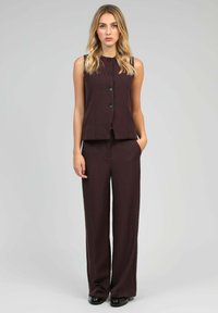 Femme se tenant debout, portant un haut sans manches boutonné marron foncé et un pantalon large assorti, avec une main dans la poche.