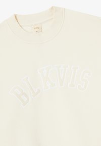 Sweter w kolorze kremowym z prążkowanym okrągłym dekoltem i białym haftowanym napisem "BLKVIS" w łuku na klatce piersiowej, rozmiar XL na metce wewnątrz dekoltu.